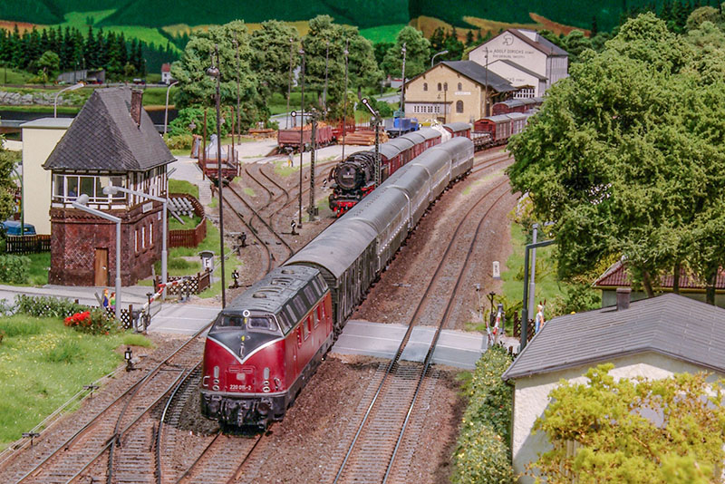 Überblick über den Bad Driburger Bahnhof. Der Eilzug nach Oberhausen hat einen Güterzug überholt und passiert den Bahnübergang an der Brunnenstraße. Links ist das Stellwerk Dwf im Bild, das Sandsteingebäude im Hintergrund ist die Güterabfertigung. Foto Stephan Rieche